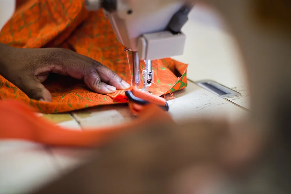 Artisan's hands guiding orange shweshwe fabric through a sewing machine